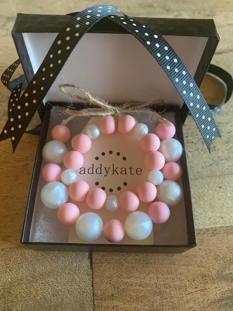 May include: A pink and white beaded bracelet set in a black gift box with white polka dots. The box is tied with a black ribbon and the bracelet is labeled "addykate".