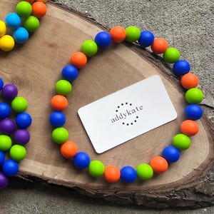 May include: A colorful beaded necklace with blue, green, and orange beads. A white business card with the text "addykate" is placed on the necklace. The necklace is displayed on a wooden surface.