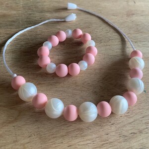 May include: A pink and white silicone necklace and bracelet set. The necklace and bracelet are made of alternating pink and white beads.