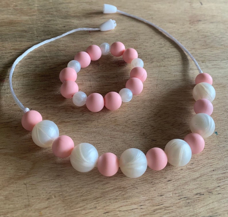 May include: A pink and white silicone necklace and bracelet set. The necklace and bracelet are made of alternating pink and white beads.
