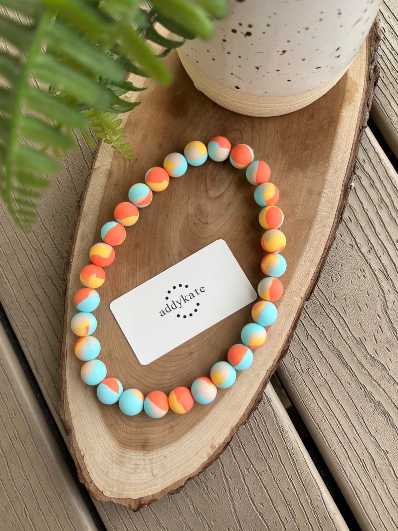 May include: A colorful beaded necklace with orange, yellow, and blue beads. The beads are arranged in a pattern of stripes and swirls. The necklace is on a wooden surface with a small card that says "addykate" on it.