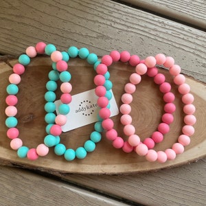 May include: Three silicone beaded bracelets in pink and turquoise colours. The bracelets are stacked on top of each other. The brand name "addykate" is printed on a white tag.
