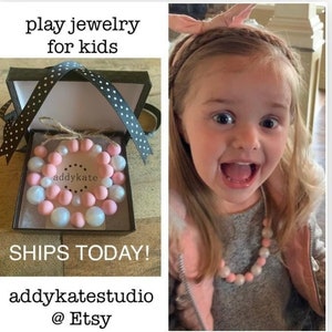 May include: A pink and white beaded necklace and bracelet set for kids. The necklace and bracelet are made of soft, colorful beads. The set is packaged in a black gift box with a white polka dot ribbon.