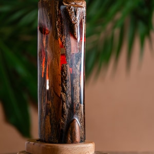 May include: A handcrafted wooden lamp with a cylindrical shape. The lamp features a combination of dark brown wood and red resin, set on a wooden base. The design highlights the natural wood grain and the warm glow of the resin.