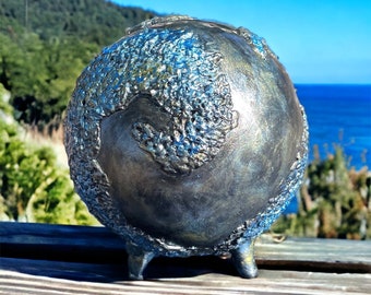 May include: A dark gray ceramic globe with a textured surface and three legs. The globe has a blue and silver iridescent finish.