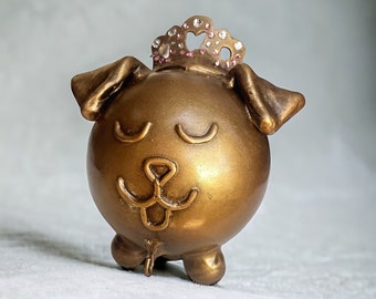 May include: A gold-colored ceramic dog figurine with a crown on its head. The dog has closed eyes and a playful expression. The crown is decorated with pink and clear rhinestones.