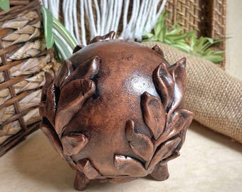 May include: A copper-colored ceramic planter with a round shape and a design of leaves around the exterior. The planter has three small feet.