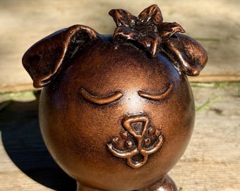 May include: A bronze-colored, spherical dog figurine with closed eyes and a flower on its head. The dog has large, floppy ears and small legs. The figurine is made of a metallic material and has a smooth, polished finish.