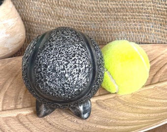 May include: A decorative, silver-colored tennis ball sculpture with a textured surface and three small feet. A bright yellow tennis ball rests beside it on a wooden leaf-shaped tray. The background features a woven texture.