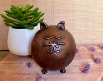 May include: A small, round, copper-colored ceramic cat figurine with a sleepy face and closed eyes. The figurine has a textured surface and is sitting on a pink surface.