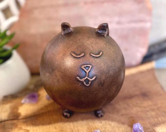 May include: A bronze-colored, spherical sculpture of a cat with closed eyes and a stylized nose and mouth. The cat sculpture has small ears and four tiny feet. The sculpture is on a wooden surface with small purple crystals.