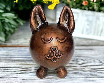 May include: A small, copper-colored ceramic figurine of a dog with closed eyes and a simple, stylized face. The figurine has a rounded body and short legs.