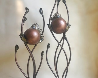 May include: Two bronze-colored decorative sculptures. Each sculpture features a spherical element held by curved, slender supports. The spheres are copper-toned, and the bases have a textured, organic appearance. The overall design is elegant and artistic.