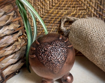 May include: A copper-colored ceramic planter with a heart-shaped design. The heart has a textured surface and is raised from the planter. The planter has three rounded legs.