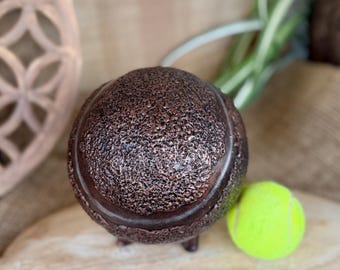 May include: A textured, bronze-colored decorative sphere with a tennis ball next to it. The sphere has a dark brown band around the middle and three small feet. The tennis ball is bright yellow-green.