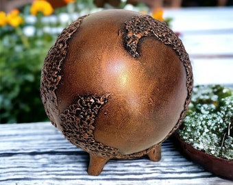 May include: A small, copper-colored sphere with a textured surface and three legs. The sphere is sitting on a wooden surface.