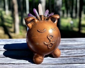 May include: A small, brown ceramic figurine of a dog with closed eyes and a smiling mouth. The figurine has a crystal crown on its head with clear and purple crystals.