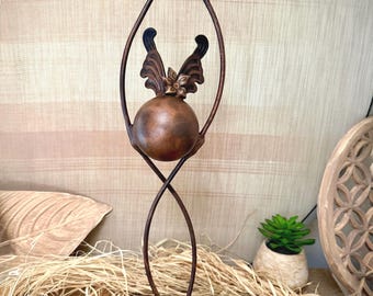 May include: A bronze-colored metal sculpture featuring a sphere with decorative butterfly-like wings and a flower. The sculpture has a curved, elongated design and sits on a textured base. The background includes a neutral-toned fabric.