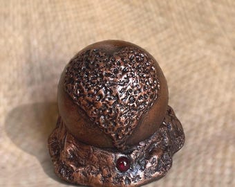 May include: A bronze-colored sculpture with a textured, heart-shaped design on top. The sculpture sits on a textured base and features a small, red gemstone. The overall aesthetic is rustic and artistic.