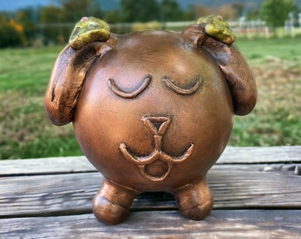 May include: A brown ceramic figurine of a dog with its eyes closed and ears covering its head. The figurine has a gold-colored glaze and a playful expression.