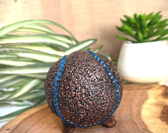 May include: A brown ceramic egg with a textured surface and a band of blue rhinestones around the middle. The egg is sitting on a wooden surface and has three small feet.