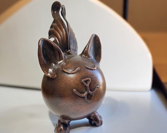 May include: A bronze-colored ceramic figurine of a cat with a spiky hairdo and a closed-eye, smiling expression.