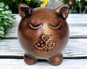 May include: A small, copper-colored ceramic cat figurine with closed eyes and a detailed nose and mouth. The cat is sitting on a wooden surface.