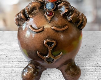 May include: A small, copper-colored figurine of a sleeping animal with a closed-eye expression. The figurine has a crown of red, blue, and clear gemstones on its head and a stylized anchor on its chest.