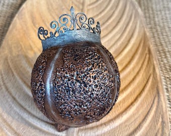 May include: A decorative, textured, bronze-colored sphere with a silver filigree crown. The sphere sits on a wooden, leaf-shaped tray. The crown adds a regal touch to the handcrafted item.