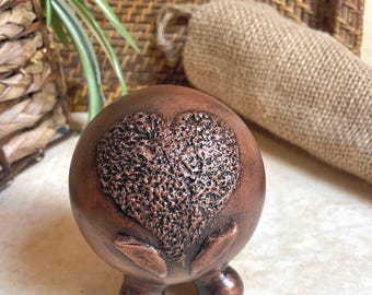 May include: A copper-colored ceramic figurine with a heart-shaped design and three legs. The heart is textured and has a raised design.