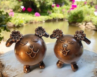 May include: Two small, bronze-colored, ceramic figurines of dogs with closed eyes and smiling expressions. Each dog has a flower-shaped design on its head.