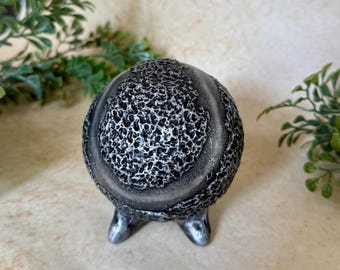 May include: A spherical object with a dark gray and black finish featuring a highly textured, porous surface. The item is supported by three small, smooth, curved feet at the base, creating a tripod-like structure for stability on flat surfaces.
