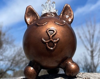 May include: A small, round, bronze-colored ceramic sculpture of a cat with closed eyes and a crown of clear quartz crystals on its head. The cat is sitting on a piece of wood.