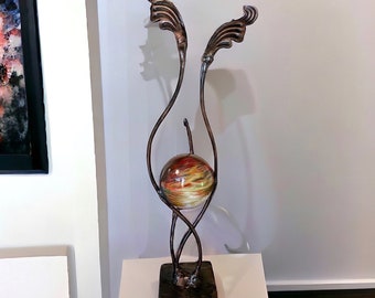 May include: A metal sculpture with a swirling design and a sphere in the center. The sphere is made of glass and has a colorful, abstract pattern. The sculpture is on a square base.