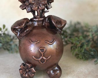 May include: A bronze-colored dog-shaped planter with a whimsical design. The planter features a smiling face with closed eyes and a small flower on one paw. Flowers sprout from the top of the dog's head, adding a decorative touch.