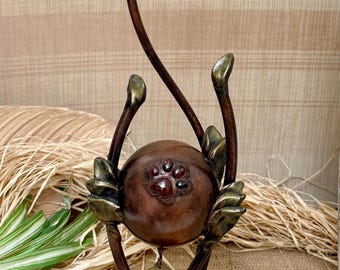 May include: A bronze-colored sculpture with a spherical center and elongated, curved stems. The sphere has embedded red and black accents. The base is textured, and a small, round tag hangs from the sphere. The sculpture has a unique, artistic design.