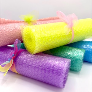 Puede incluir: Una colección de rollos de plástico de burbujas en colores vibrantes que incluyen amarillo neón, rosa claro, morado, verde y azul. Cada rollo presenta un patrón repetitivo de burbujas en forma de corazón y está atado con un lazo transparente.