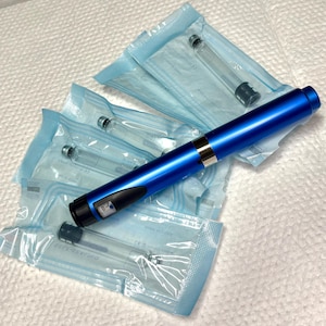 May include: A blue medical pen with a silver band and black accents rests on top of several sealed, clear glass vials. The vials are in light blue packaging. The pen has a digital display.