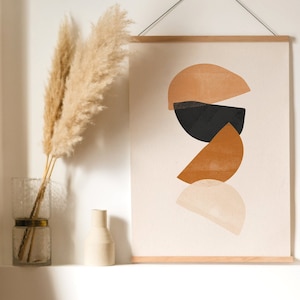 May include: A minimalist abstract art print with geometric shapes in shades of brown, beige, and black. The print is framed in a light wood frame and hung on a white wall. A small framed print with a sunburst design is to the right of the larger print.