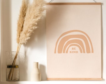 May include: A beige wall with a framed print of a rainbow with the text "Be Kind" in brown. A smaller framed print of a sunset landscape is to the right of the larger print. A vase with dried pampas grass is on a shelf below the prints.