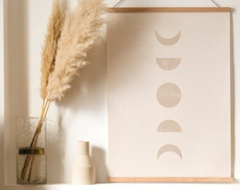 May include: A beige framed print with a minimalist design of a moon phases illustration. A smaller framed print with a sunset landscape in shades of pink and orange hangs below. A vase with dried pampas grass sits on a shelf.