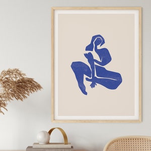 May include: A framed art print featuring a minimalist design of a seated figure in a vibrant blue hue against a cream background. The artwork is displayed in a light wood frame, adding a touch of warmth to the modern aesthetic.