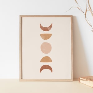 May include: A framed print with a minimalist design featuring a vertical arrangement of five abstract shapes in shades of brown, orange, and pink, resembling moons or crescents. The print is set against a white background and framed in a light wood frame.