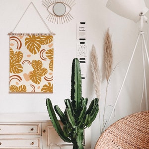 May include: A white wall with a wooden frame tapestry featuring a pattern of brown and orange tropical leaves and pink and orange abstract shapes. The tapestry is hanging on a white wall with a white lamp and a potted cactus plant in front of it.