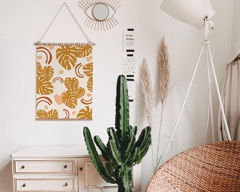 May include: A white wall with a wooden frame tapestry featuring a pattern of brown and orange tropical leaves and pink and orange abstract shapes. The tapestry is hanging on a white wall with a white lamp and a potted cactus plant in front of it.
