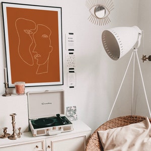 May include: A white record player with a brown record on the turntable. The record player is sitting on a white dresser with a candle holder and candles. There is a framed print of a line drawing of two faces on the wall behind the record player. The print has a burnt orange background.