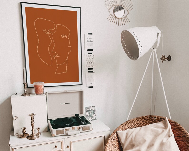May include: A white record player with a brown record on the turntable. The record player is sitting on a white dresser with a candle holder and candles. There is a framed print of a line drawing of two faces on the wall behind the record player. The print has a burnt orange background.