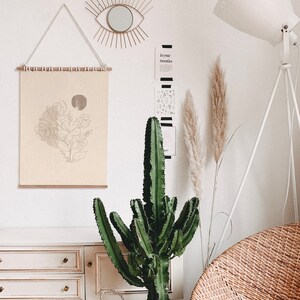 May include: A white dresser with three drawers, a cactus plant, a white floor lamp with a tripod base, and a beige wicker chair. A framed print with a line drawing of flowers hangs on the wall above the dresser. The wall also has a gold sunburst mirror and a black and white print with the text "In your twenties".