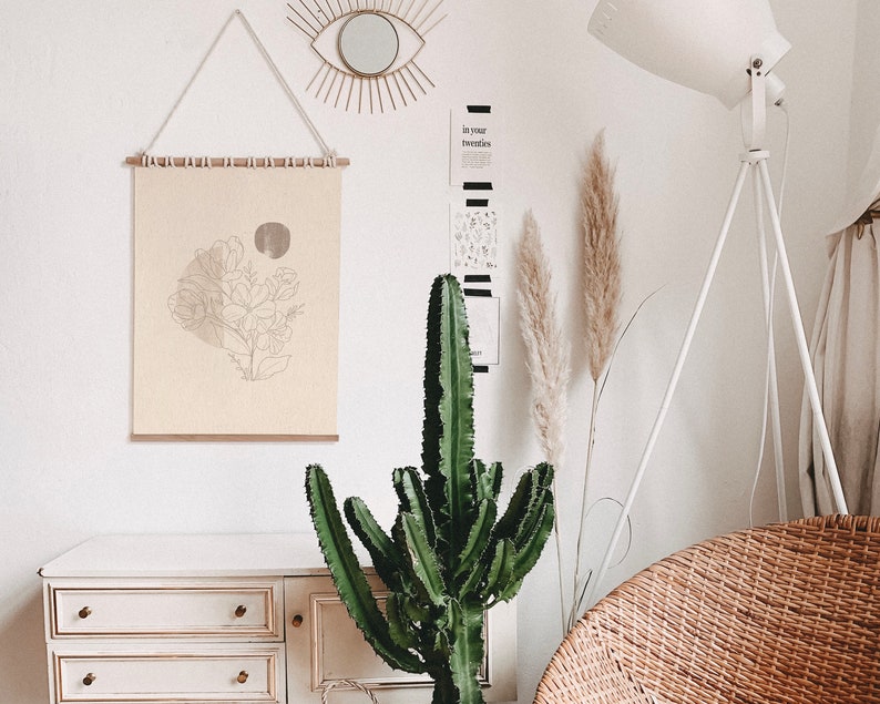 May include: A white dresser with three drawers, a cactus plant, a white floor lamp with a tripod base, and a beige wicker chair. A framed print with a line drawing of flowers hangs on the wall above the dresser. The wall also has a gold sunburst mirror and a black and white print with the text "In your twenties".