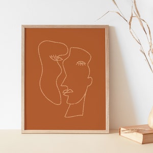 May include: A framed print with a minimalist line drawing of two faces in white on a burnt orange background.